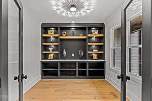A modern home office features built-in black shelves with wooden accents, decorative items, and framed photos. Double glass doors and large windows let in natural light. A contemporary chandelier hangs from the ceiling.