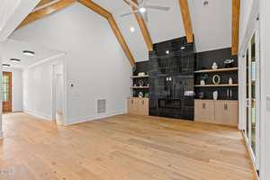 A spacious living room with light wood flooring, vaulted ceiling with exposed wood beams, and a black tiled fireplace flanked by built-in shelves and cabinets. Sliding glass doors are on the right wall.