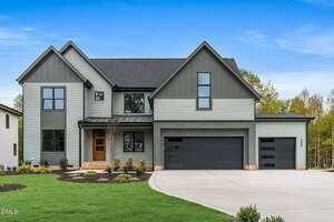 A modern two-story house at 3906 Cedar Knolls Drive, Youngsville, with gray siding, dark trim, large windows, and three garage doors. The front yard has green grass, young trees, and a wide driveway under a clear blue sky.