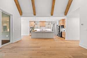 Spacious modern kitchen with light wood floors, exposed ceiling beams, center island with pendant lights, light wood cabinetry, stainless steel appliances, and large glass door leading to another room.