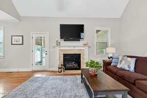 A living room with a wall-mounted TV above a fireplace, a coffee table with a plant, a maroon sofa with pillows, a rug, a lamp, and windows letting in natural light.