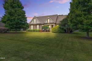 A single-story house with gabled roof and covered porch is surrounded by neatly trimmed bushes and large trees, set on a spacious, well-maintained lawn at dusk.