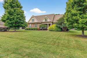 A single-story brick house with gabled roof, front porch, and manicured shrubs sits on a large grassy lawn bordered by mature trees under a blue sky with scattered clouds.