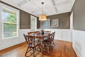 A dining room with a round wooden table and six matching chairs, a light fixture above, hardwood floors, coffered ceiling, two framed pictures on the wall, and large windows with blinds letting in natural light.