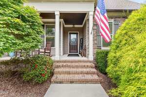 Brick steps lead to a covered front porch with a wooden door, rocking chairs, a "Welcome" sign, potted plants, and an American flag. Green shrubs and trees line the walkway.