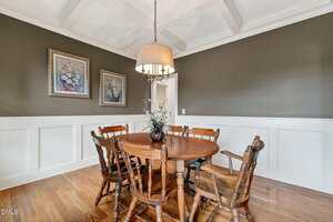 A dining room with a round wooden table, six matching wooden chairs, a hanging light fixture, coffered ceiling, dark olive green upper walls, white wainscoting, and two framed floral paintings.