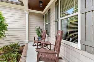 Two wooden rocking chairs with a small side table sit on a covered porch with beige siding, green shutters, brick trim, and potted greenery near a large window.
