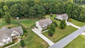 Aerial view of a suburban neighborhood with three single-family homes, large front and backyards, driveways, landscaped lawns, and a wooded area behind the houses. A street runs in front of the properties.