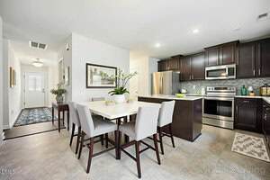 A modern kitchen and dining area with dark wood cabinets, stainless steel appliances, a white dining table with six upholstered chairs, and decorative plants. The space is well-lit and features light-colored flooring.