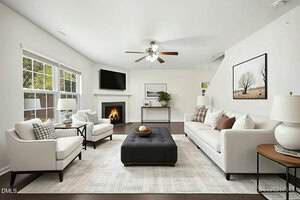 A bright living room with white walls, two white sofas, two armchairs, a black tufted ottoman, fireplace, flat-screen TV, large windows, wall art, a plant, and a ceiling fan.