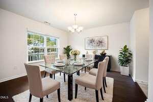 A dining room with a glass table set for six, beige upholstered chairs, a white rug, dark wood floors, white walls, potted plants, a sideboard, and a chandelier. Large window lets in natural light.