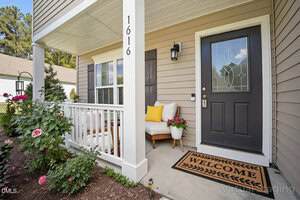 A porch with beige siding, black front door with glass panel, "Welcome" doormat, chair with yellow and white cushions, small white table, and potted pink flowers next to a garden bed. House number 1616 is displayed on a column.