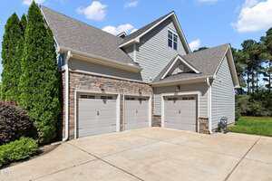 A three-car garage attached to a modern house with gray siding, stone accents, and a concrete driveway. Bushes and trees are visible on the left and right sides, with a green lawn in the background.