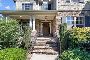 A stone walkway with steps leads to a covered front porch of a house with a dark brown door, sidelights, two white pillars, and stone and shingle exterior walls. Shrubs and bushes line the entrance.