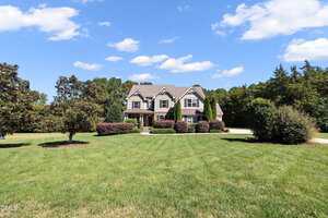 A two-story house with a covered porch sits on a large, well-maintained lawn. The house features gables, dormer windows, and is surrounded by shrubs and trees under a blue sky with clouds.