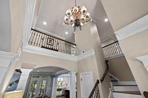 A view of the two-story home at 690 Hawthorne Place features a staircase, balcony with black railings, beige walls, white trim, and a decorative chandelier. The kitchen and large windows are visible in the background.