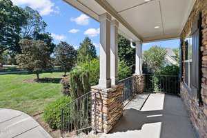 A covered front porch with stone pillars and metal railings overlooks a landscaped yard with green grass, shrubs, and trees under a blue sky with scattered clouds.