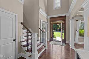 A spacious entryway with hardwood floors, a wooden staircase with white trim and black balusters, tall ceilings, and large windows allowing natural light. An open front door leads to a green lawn outside.