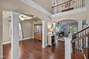 Open foyer with hardwood floors, white columns, and crown molding. A wooden staircase with a black railing leads upstairs. Dining and living rooms are visible, both furnished with wood pieces and neutral decor. Natural light fills the space.