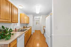 A galley kitchen with wood cabinets, laminate countertops, white appliances, and wood-look flooring. A small window is above the sink, and a green plant sits on the counter. The kitchen leads to a door at the far end.