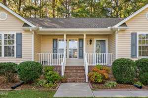 A beige single-story house with blue shutters, a covered front porch, white railings, brick steps, and neatly trimmed bushes and plants in the landscaped front yard.