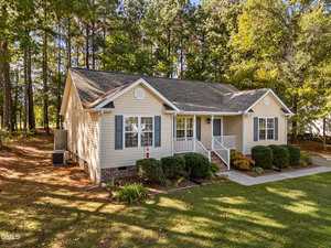 Single-story beige house with blue shutters, white trim, and a covered front porch. Surrounded by trees and shrubs, the home features a concrete driveway and a neatly maintained front lawn.