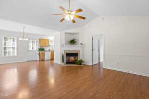 A spacious living area with wood flooring, a ceiling fan with lights, a white fireplace, and an open view into a kitchen with light wood cabinets. Large windows provide natural light.