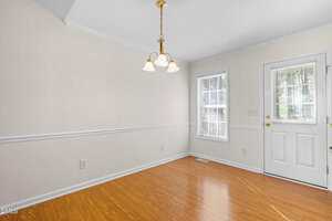 A small, empty room with light beige walls, wood flooring, a white door with glass panes, two windows with blinds, and a ceiling light fixture with three bulbs.
