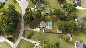 Aerial view of a suburban home with a fenced backyard, patio, and pool. The property is surrounded by trees and lawns, with neighboring houses and curved roads visible in a residential neighborhood.