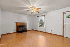 A living room with light wood flooring, a white ceiling with a ceiling fan, a fireplace with a wood mantel, a window with white curtains, and a white door with a gold handle.