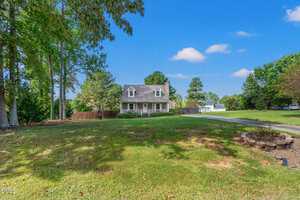 A two-story house with a front porch sits on a large grassy yard with scattered trees. A curved driveway leads to the house, and there is a fire pit area in the foreground. The sky is clear with a few clouds.