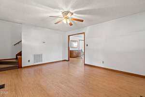 A spacious, empty living room with light-colored walls, wood laminate flooring, a ceiling fan with lights, and an open doorway leading to a dining area. Carpeted stairs are visible on the left side of the image.