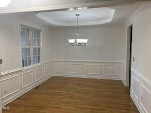 A dining room with white wainscoting, hardwood floors, a chandelier, a tray ceiling, and a large window on the left wall. The room is empty and has neutral-colored walls.