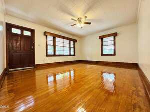 Empty living room with polished hardwood floors, white walls, a dark wooden front door, three windows with wooden trim, and a ceiling fan with lights. Natural light fills the space.