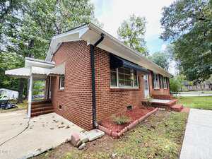 A one-story red brick house with white trim, black shutters, and a white awning over the side entrance. The yard has red mulch, small shrubs, and is surrounded by mature trees. A concrete walkway leads to the front steps.