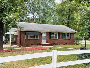 A single-story red brick house with a gray roof, white trim around the windows and door, and a covered side entrance. The yard features grass, red mulch, and a white wooden fence in the foreground.