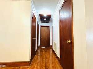 A hallway with polished wooden floors, white walls, three closed wooden doors on either side, and a closed door at the end; a ceiling light fixture is on, and an air vent is visible on the right wall.