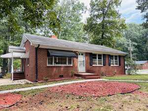 Single-story red brick house with black shutters and a white front door, set among trees. There is a small covered side entrance and a yard with red mulch and sparse grass. A residential street is visible in the background.