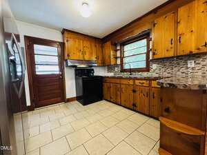 A kitchen with wooden cabinets, a black stove and range hood, mosaic tile backsplash, tile flooring, a stainless steel refrigerator, a window above the sink, and a wooden door with glass panels.