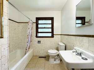 A bathroom with a bathtub and floral shower curtain, toilet, wall-mounted sink with faucet, mirror, tiled walls and floor, and two windows with wooden trim.