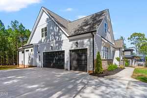 A modern two-story house with white siding, black garage doors, multiple windows, a gabled roof, and a wide concrete driveway, surrounded by trees and landscaping.