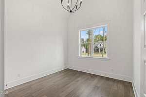 A small, empty room with white walls, a wood floor, a modern black chandelier, and a window showing a view of houses and trees outside.
