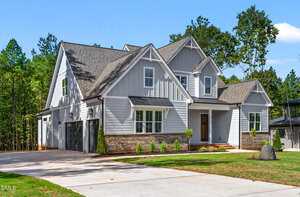 A two-story modern suburban house with gray siding, white trim, brick accents, a covered front porch, and a double garage. The yard has fresh landscaping and a driveway, with trees in the background under a clear sky.