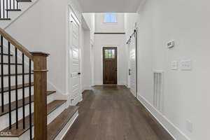 A view of a modern entryway with dark wood flooring, white walls, a wooden front door with glass panels, multiple white doors, a staircase with wooden steps and black railing, and a high ceiling with a window.