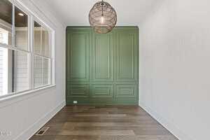 A narrow room with light-colored walls, a large green paneled accent wall at the end, wood flooring, a window on the left, and a round pendant light hanging from the ceiling.