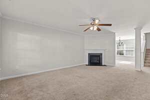 A spacious living room with beige carpet, a ceiling fan with lights, a white fireplace in the corner, light gray walls, and an open view into a dining area with stairs leading up on the right side.