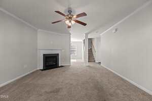 A carpeted living room with beige walls, a ceiling fan with lights, a white fireplace, a window at the back, and a staircase leading upstairs. The room is unfurnished and has white trim.