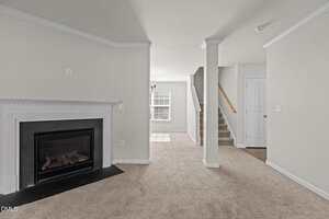 A room with beige carpet, a white fireplace on the left, a staircase with a wooden handrail on the right, and a window in the background letting in natural light. Walls and trim are painted white.