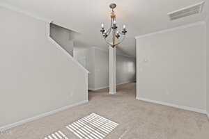 A room with beige carpet, white walls, and a modern black chandelier. An angled staircase with a partial wall leads upward. Sunlight casts window shadows on the floor. Visible door and air vent on the ceiling.