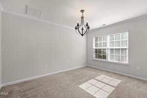 Empty room with beige carpet, light gray walls, white trim, a black metal chandelier on the ceiling, and a large window with white blinds letting in natural light.